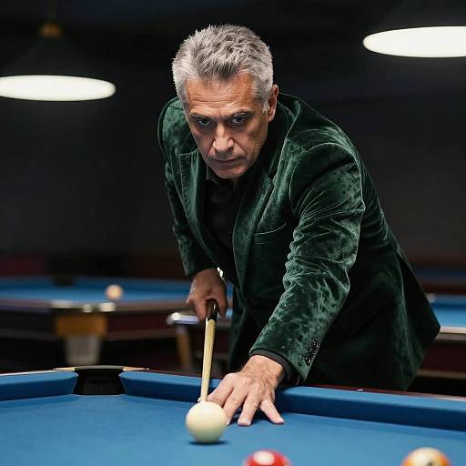 Photograph of an intense, gray-haired middle-aged man in a dark green velvet jacket, leaning over a blue pool table, aiming a cue stick at