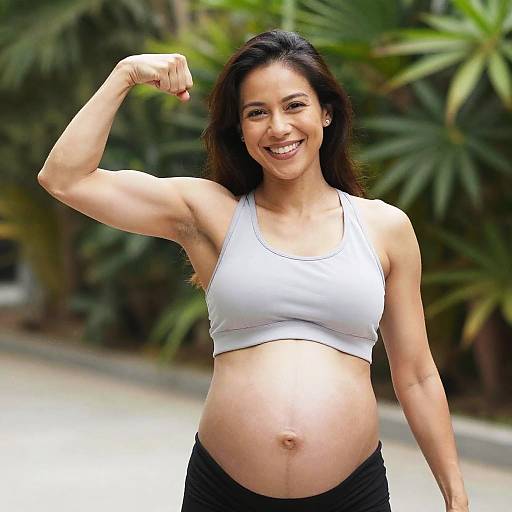 Pregnant woman with brown hair, smiling, flexing right arm, wearing white sports bra, black pants, green leafy background, photograph.