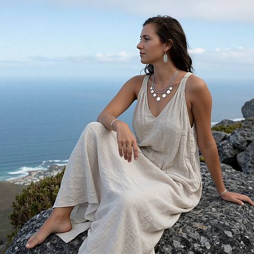 Photograph of a dark-haired woman in a white, sleeveless dress and white necklace, sitting on a rocky cliff overlooking a blue ocean and cloudy sky