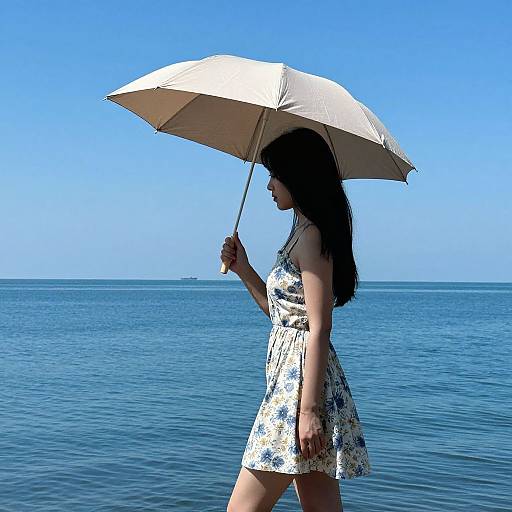 Photograph of a woman with long dark hair, wearing a white floral dress, holding a white umbrella, walking on a blue ocean shore under a clear