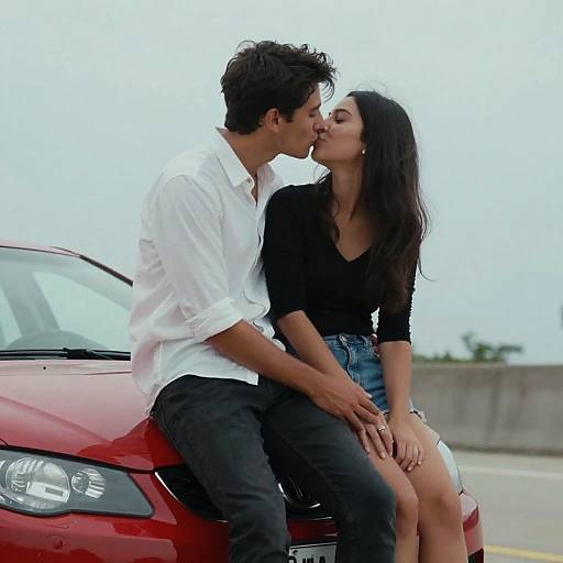 Photograph of a young couple kissing passionately while sitting on the hood of a red car, wearing white shirt and black top, blue shorts, in an