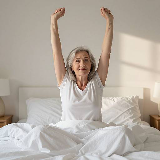 Photograph of an elderly woman with gray hair, wearing a white t-shirt, stretching with arms raised, sitting in a sunlit bed with white pillows