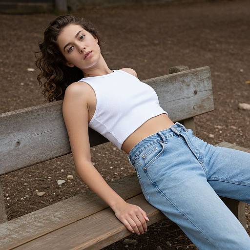 Photograph of a young woman with wavy brown hair, wearing a white crop top and light blue jeans, reclining on a wooden bench in a