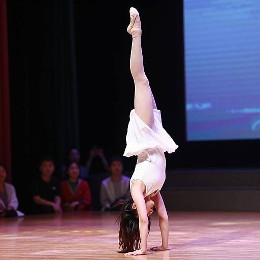 Photograph of a female gymnast performing a handstand with one leg raised high, wearing a white leotard, on a brightly lit stage.