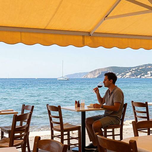 Photograph of a man in a gray shirt and shorts, sipping from a cup, seated at a beachside table under a yellow canopy with a