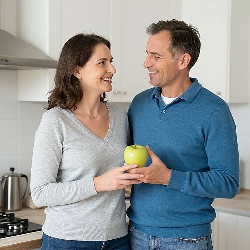 Photograph of smiling couple in a bright kitchen; woman in gray sweater, man in blue sweater, both holding a green apple.