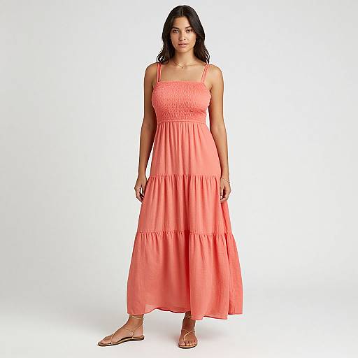 Photograph of a dark-haired woman in a coral, sleeveless, tiered maxi dress with thin straps, standing against a white background. She wears