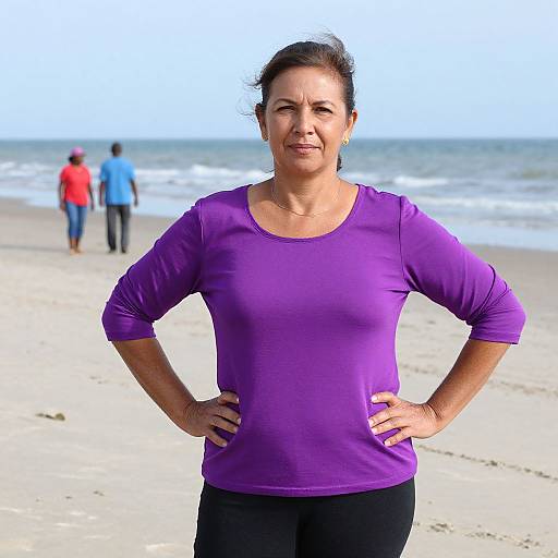 Photograph of a middle-aged woman with dark hair in a purple long-sleeve shirt and black pants, standing on a beach with two people in
