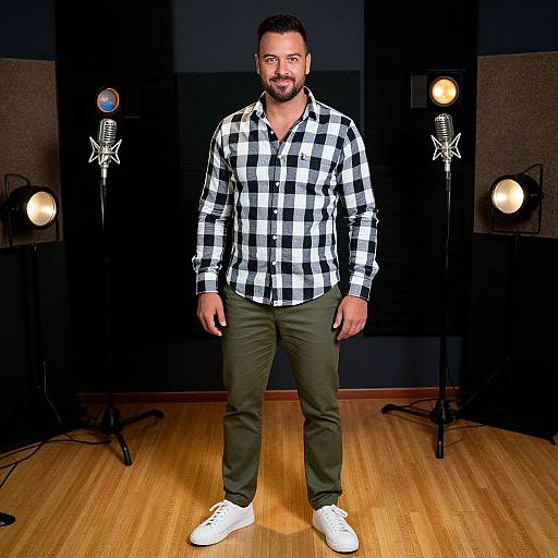 Photograph of a bearded man with short dark hair, wearing a black-and-white checkered shirt, olive-green pants, and white sneakers, standing