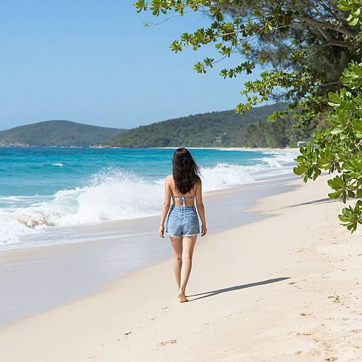 Photograph of a woman with long black hair in a blue denim romper walking on a sunny, tropical beach with turquoise waves and green hills in the