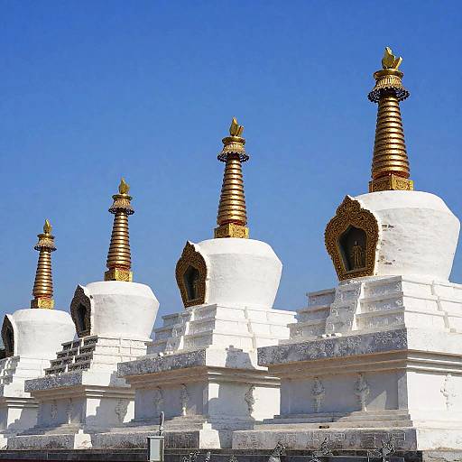 Photograph of four white Buddhist stupas with golden-tipped spires, set against a clear blue sky, showcasing traditional Tibetan architecture.