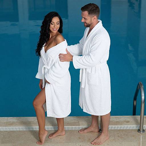 Photograph of a smiling couple in white bathrobes standing by a blue pool, with the man gently touching the woman's waist.