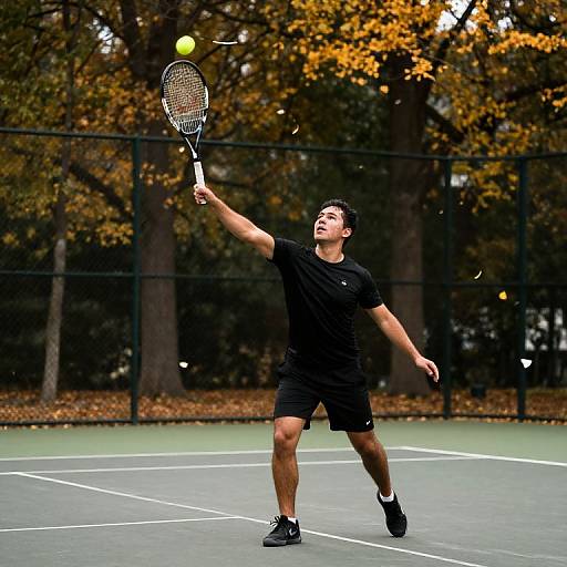 Photograph of a muscular male tennis player in black attire, mid-serve, with a yellow tennis ball and racket, on an outdoor court with autumn