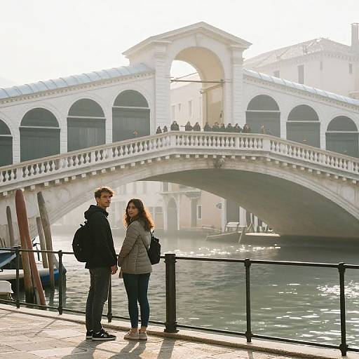 Photograph of a couple standing on a Venice bridge, gazing at the canal with a white arched bridge in the background.