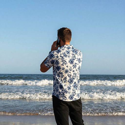 Photograph of a man with short brown hair, wearing a white floral shirt and black pants, standing at the beach, talking on a cellphone, with