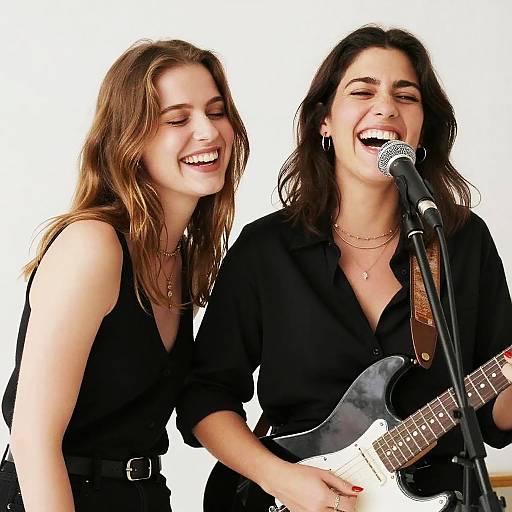Photograph of two smiling women with long brown hair, wearing black tops, singing and playing guitar on stage with microphone.