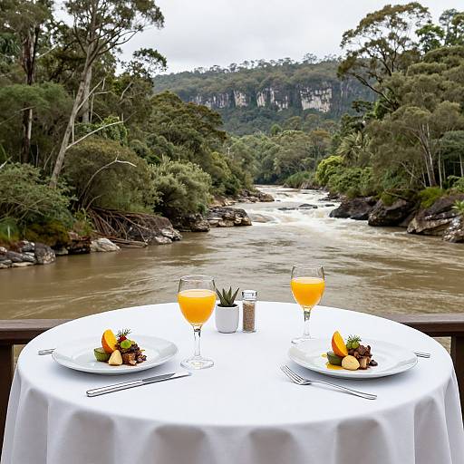 Romantic riverside table setting with two plates of fruit and orange drinks, overlooking a lush forest and flowing river.