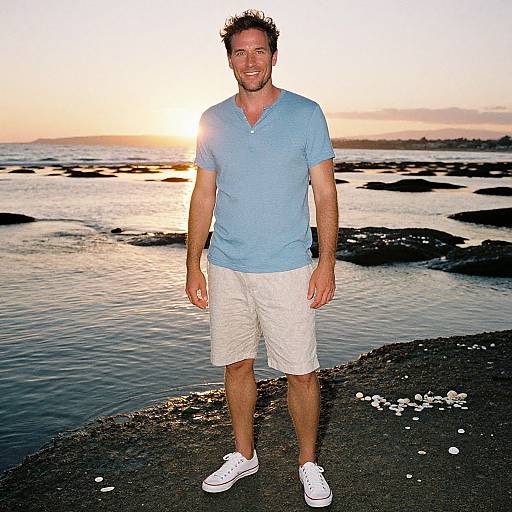 Photograph of a smiling man with short brown hair, blue shirt, white shorts, and white sneakers standing on a rocky shoreline at sunset.
