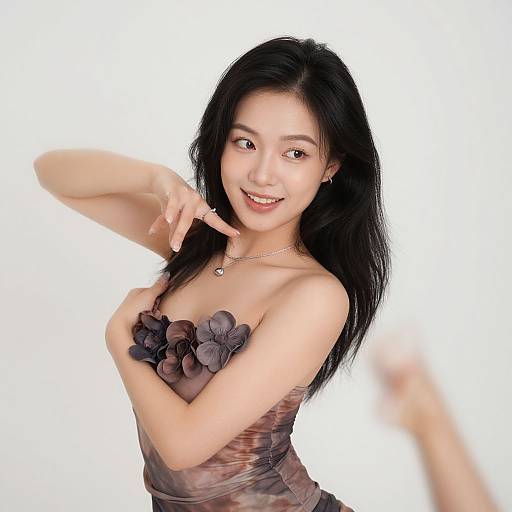 Photograph of a smiling Asian woman with long black hair, wearing a strapless brown dress with black flower embellishments, pointing playfully with her right