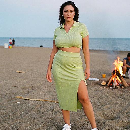 Photograph of a curvy woman with dark hair in a light green, ribbed, two-piece outfit with a high slit, standing on a sandy