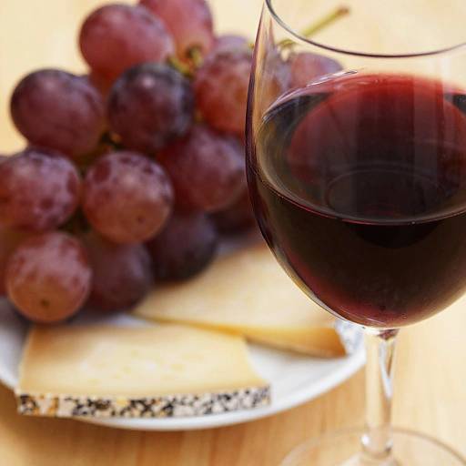 Photograph of a glass of red wine beside a plate of grapes and cheese on a wooden table.