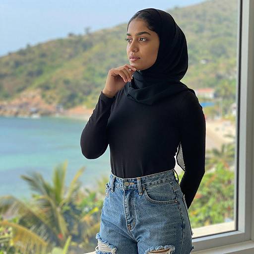 Photograph of a young Black woman with a black hijab, wearing a black long-sleeve top and high-waisted, ripped blue jeans