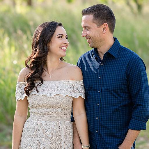 Photograph of a smiling couple standing outdoors; woman in white lace off-shoulder dress, man in navy checkered shirt, green blurred background.