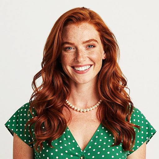 Photograph of a smiling woman with long red hair, wearing a green polka-dot dress and pearl necklace, against a white background.