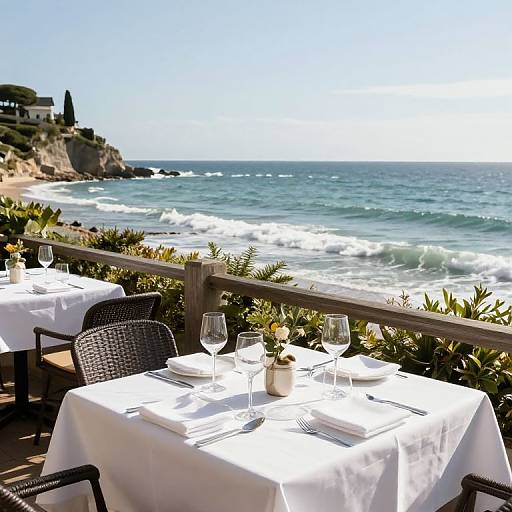 Seaside restaurant patio photograph: white tablecloths, wicker chairs, wine glasses, ocean view with waves, sunny sky, and coastal greenery