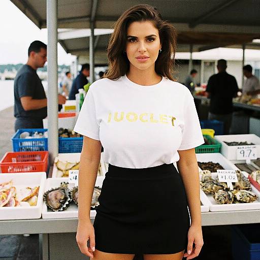 Photograph of a brunette woman with wavy hair, wearing a white Hooters shirt and black skirt, standing at a seafood market stall. Background