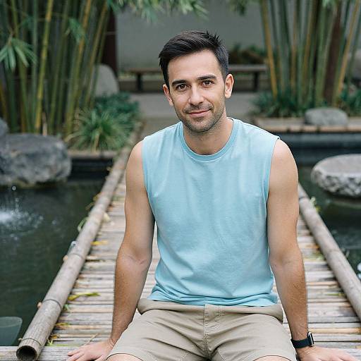 Photograph of a smiling, dark-haired man with a trimmed beard, wearing a light blue sleeveless shirt and beige pants, sitting on a bamboo bridge