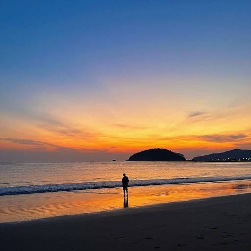 Silhouette of a person standing on a beach at sunset, reflecting vibrant orange and blue sky, with distant hills on the horizon. Photograph.
