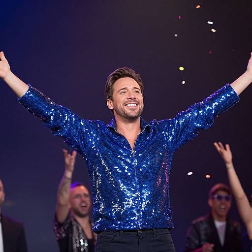 Photograph of a smiling man with short brown hair, wearing a sequined blue shirt, arms raised, on stage with cheering background.