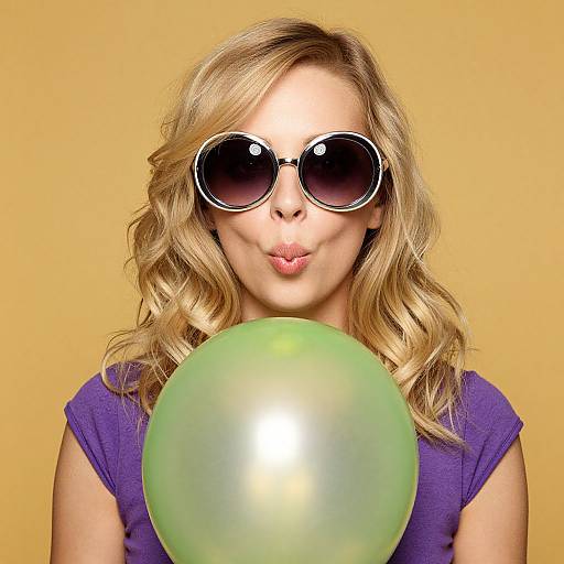 Photograph of a blonde woman with wavy hair, wearing oversized black sunglasses and a purple shirt, blowing a kiss while holding a green balloon against a