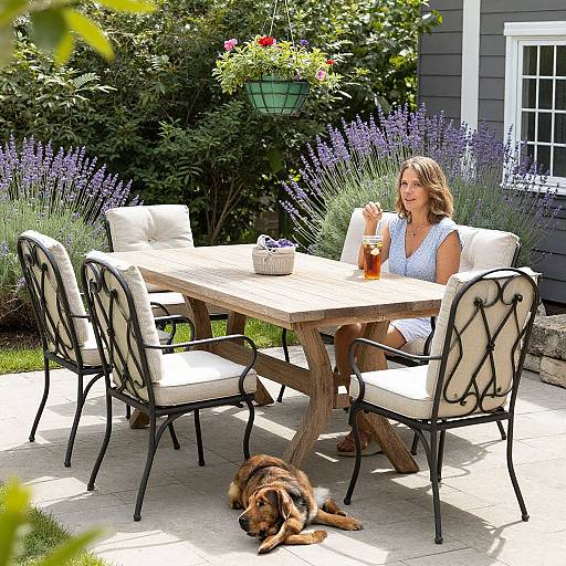 Photograph of a smiling woman in a white dress, sitting at a wooden outdoor table with black wrought iron chairs, sipping iced tea, surrounded