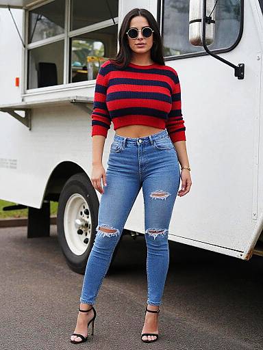 Photograph of a woman with long dark hair, wearing a red and black striped sweater, ripped blue jeans, and black heels, standing in front of