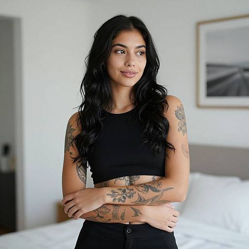 Photograph of a tattooed woman with long black hair, wearing a black crop top, standing in a bright, minimalist bedroom.
