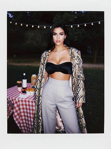 Photograph of a woman with dark hair, wearing a snake-print coat, black strapless top, and high-waisted white pants, standing in
