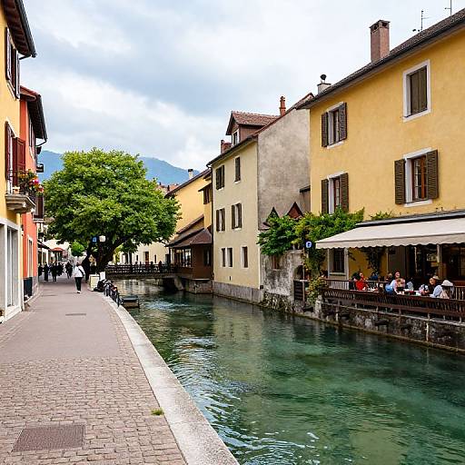 Photograph of a quaint European canal with yellow and beige buildings, people dining under a white awning, and a green tree lining the cobblestone