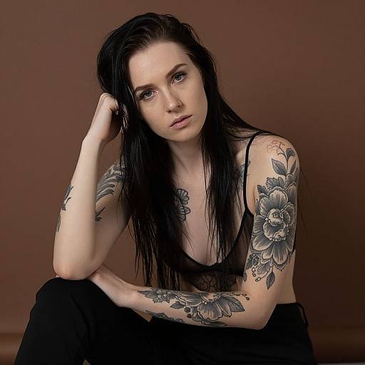 Photograph of a pale-skinned, black-haired woman with intricate floral tattoos on her arms, sitting against a brown background, wearing a black tank top