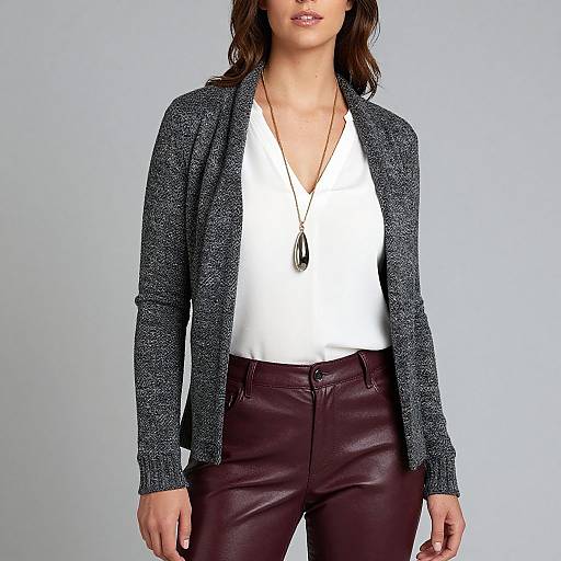 Photograph of a woman with wavy brown hair, wearing a gray textured cardigan, white V-neck blouse, and maroon leather pants, against