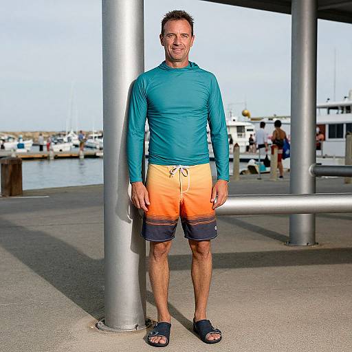 Photograph of a smiling middle-aged man with short dark hair, wearing a teal long-sleeve shirt, orange shorts, black sandals, standing under