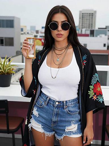 Photograph of a dark-haired woman with round sunglasses, wearing a white tank top, distressed denim shorts, and a black floral kimono, holding a