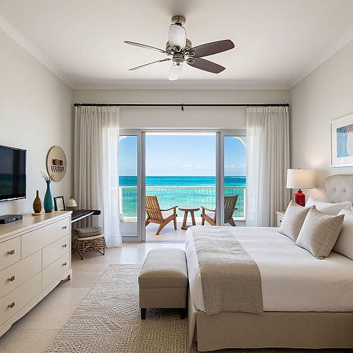 Bright beachside bedroom photograph: white bed with pillows, gray blanket, ceiling fan, large window with ocean view, white dresser, wooden chairs.