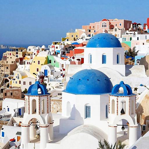 Photograph of a vibrant, sunlit Mediterranean village featuring white buildings with bright blue domes and roofs, surrounded by multicolored structures under a clear