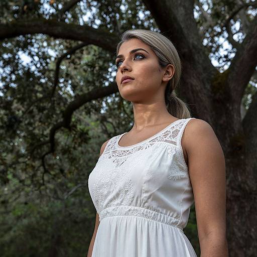 Photograph of a young woman with fair skin and blonde hair, wearing a white lace sleeveless dress, standing in a forest with dark, leafy