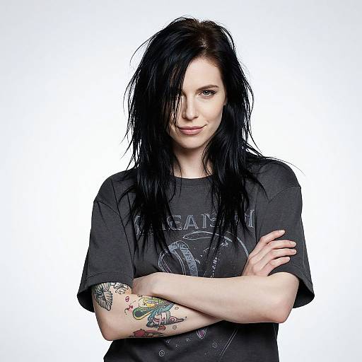 Photograph of a pale-skinned woman with black, wet-looking hair, wearing a black graphic t-shirt, arms crossed, displaying colorful tattoos on her