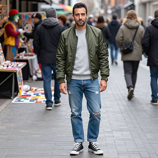 Photograph of a bearded man with dark hair, wearing a green jacket, white tee, ripped jeans, and black sneakers, standing on a busy