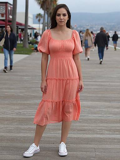 Photograph of a young woman with long dark hair, wearing a coral pink dress and white sneakers, standing on a coastal boardwalk with blurred pedestrians and