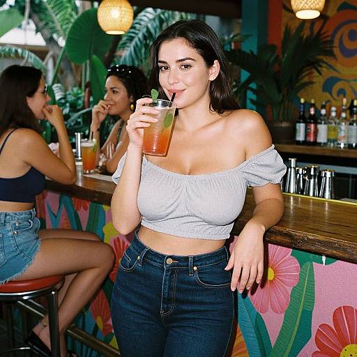 Photograph of a young woman with light skin and dark hair, sipping a green drink at a vibrant bar with floral-patterned decor, wearing a